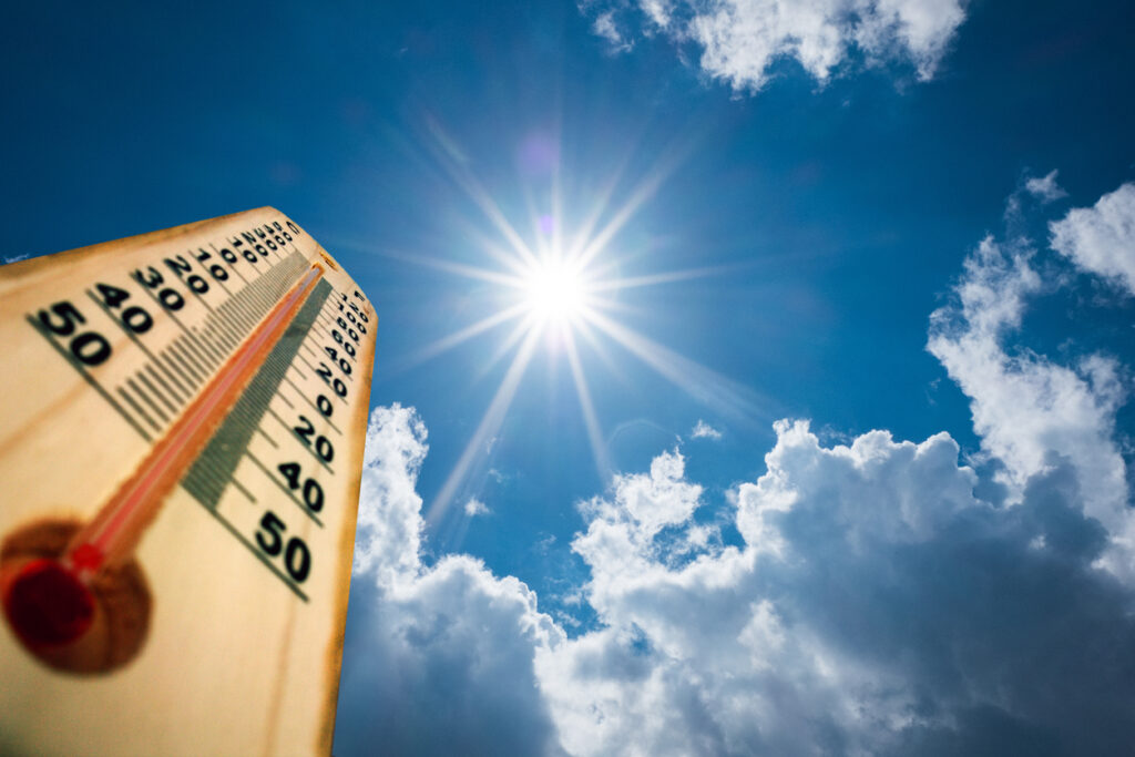 A close up of a thermometer on a hot summer day set against the background of the sun in the sky.