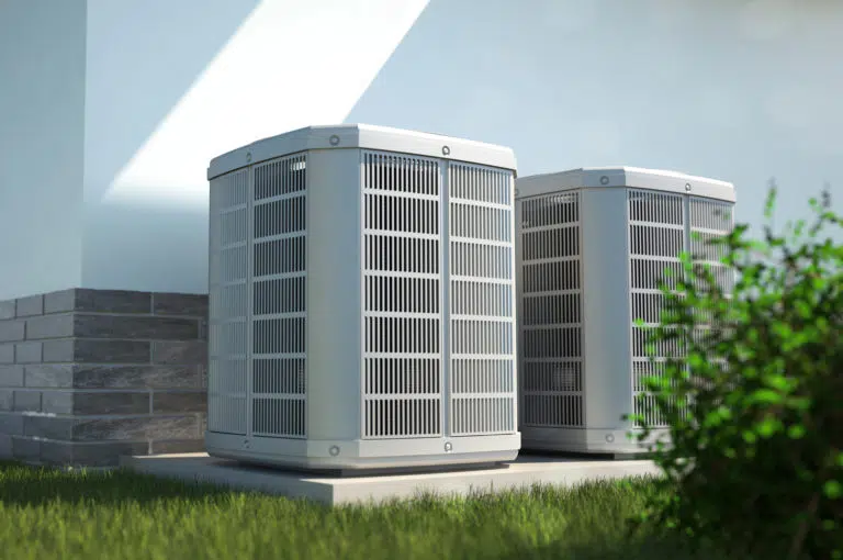 Two HVAC units outside a white stucco home, with small green bush in foreground, to the right.