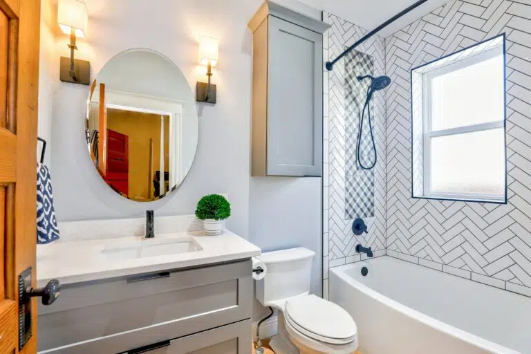A modern, white bathroom with a single vanity and a combo shower and bathtub. Herringbone-style white tiles around tub. Round mirror above sink.